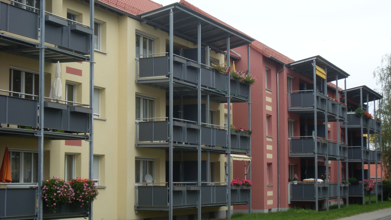 Das Nachher-Bild eines Gebäudes in der Gerhart-Hauptmann-Straße 1-11. Moderne Balkone in dunklem Grau wurden hinzugefügt, während das Gebäude in einem Pastellgelb und -rot erstrahlt.