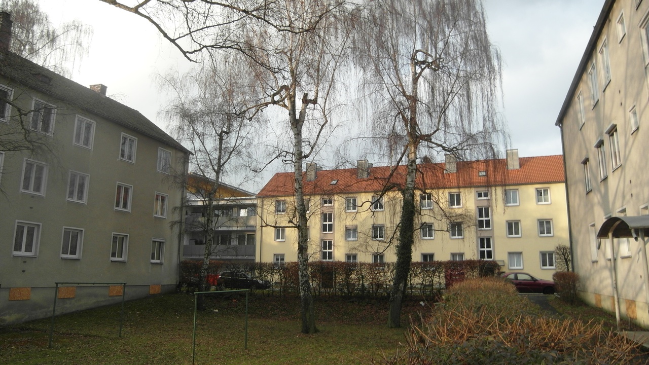 Reihe von drei Häusern in der Gerhart-Hauptmann-Straße 2-16, mit einem Baum in der Mitte. Das linke Haus besitzt eine grüne Grundierung, das mittlere eine gelbe und das rechte eine beige. Die Gebäude tragen die Spuren der Zeit, und die Hausfarben sind verschmutzt.