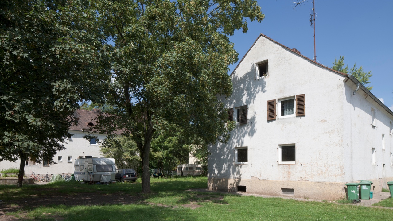 Ein Vorher-Bild des Gebäudes in der Herderstraße 1 zeigt eine ältere Struktur, bei der der weiße Putz an einigen Stellen abgebrochen ist.